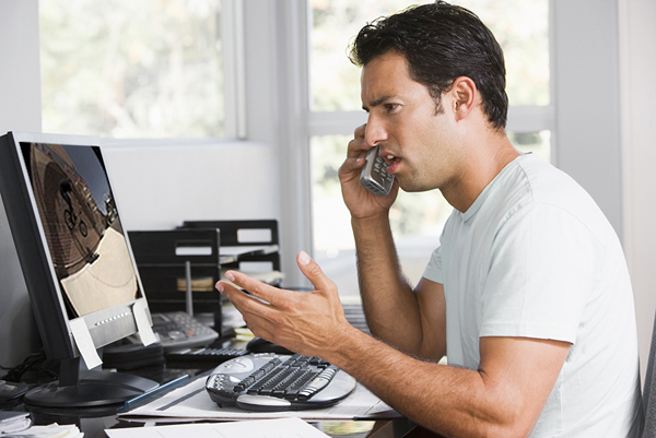 Man in home office on telephone using computer and frowning